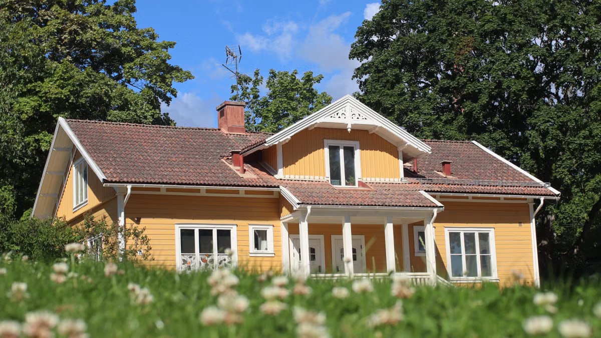 A picturesque yellow bungalow with a tiled roof surrounded by lush greenery under a bright blue sky.